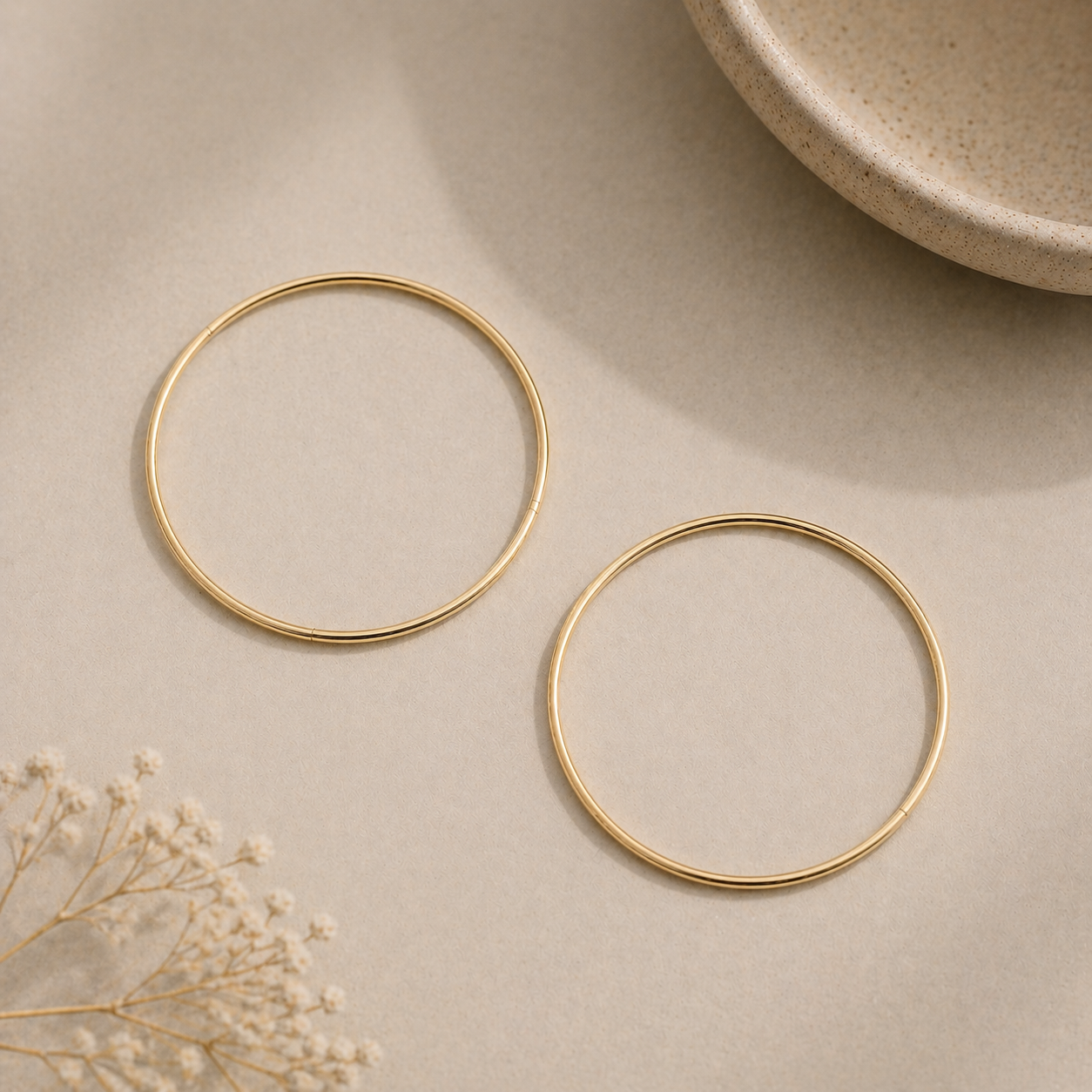 Gold hoop earrings on a beige surface with a ceramic plate and dried flowers.